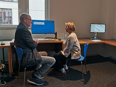 a man and a woman sitting facing each other in front of a pc screen