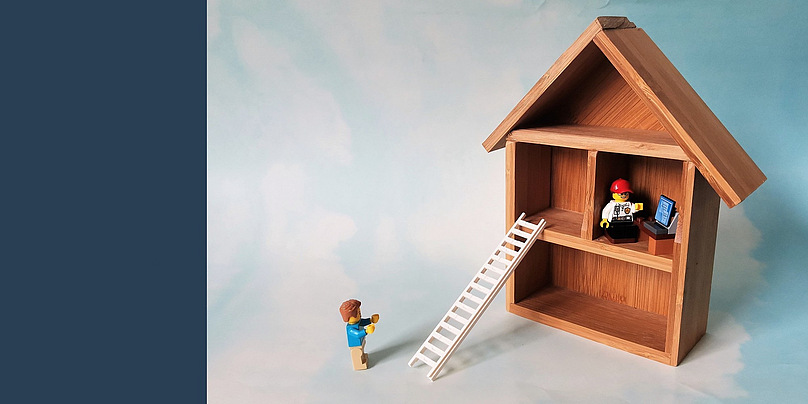 Eine Legofigur sitzt im ersten Stock eines Setzkastenhäusches aus Holz. Die Figur sitzt vor einem Computer. An das Haus ist eine Leiter gelehnt. Am Fuß der Leiter steht eine weitere Legofigur mit erhobenen Armen.