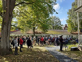 Gäste im Garten des Hauses für Barrierefreiheit. Die meisten stehen, einige Rollstuhlfahrer. Ein Kameramann filmt.
