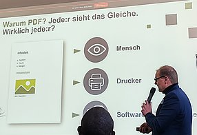 Markus Erle steht im Profil mit einem Mikro in der Hand vor seiner Präsentation, die an die Wand projiziert wird.