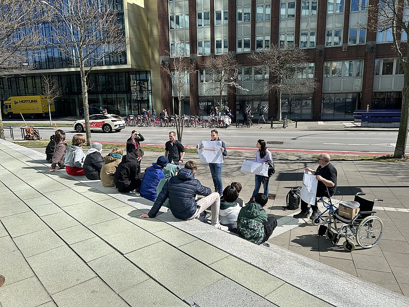 Gruppe von Jungen sitzt draußen auf Treppe und hört Bezirksamtsmitarbeitern bei einer Erklärung zu. Die Erwachsenen halten Straßenpläne hoch.