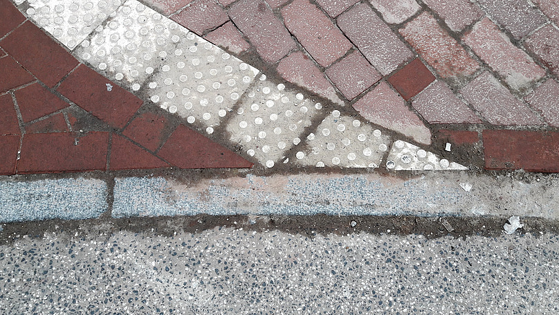 Ausschnitt einer Straßenszene: verschiedene Bodenbeläge (Asphalt und Backsteine) und Noppensteine, von oben fotografiert