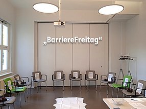 Foto mit Blick in den Schulungsraum, Stuhlkreis mit 10 Stühlen. Im Hintergrund der Schriftzug: BarriereFreitag
