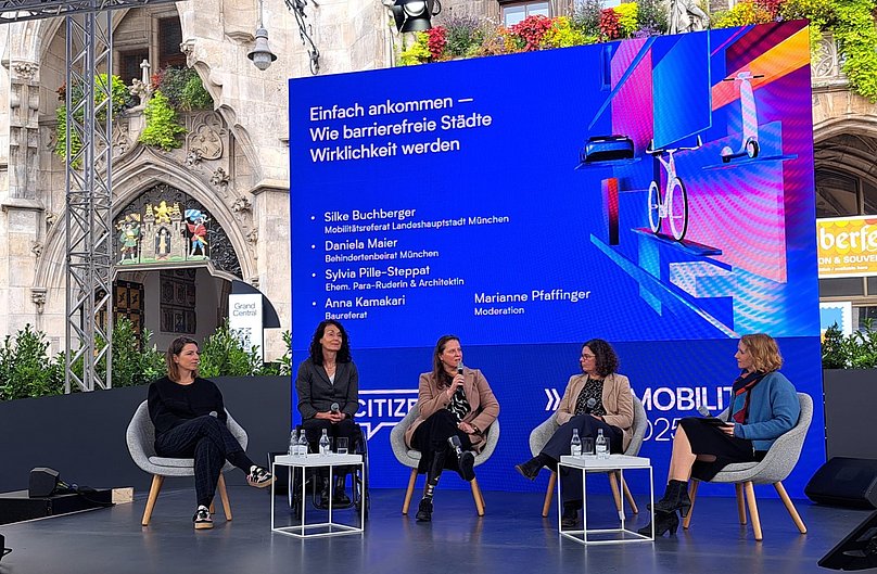 Eine Gruppe Frauen sitzt im Halbkreis auf einer Bühne, eine davon im Rollstuhl. Im Hintergrund ist das Programm auf einer Leinwand zu sehen.