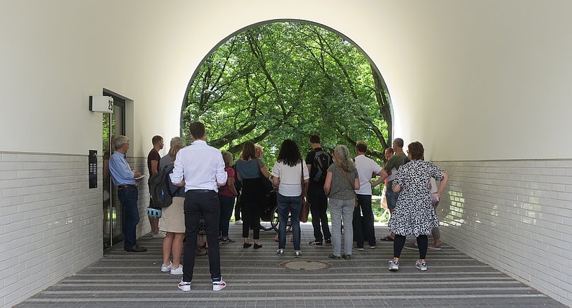 Eine Gruppe von hinten, die in einem Häuserdurchgang steht, der sich bogenförmig zu einer Grünfläche hin öffnet.