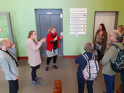 Eine Gruppe Menschen hört einer Frau zu, neben ihr steht eine DGS-Dolmetscherin